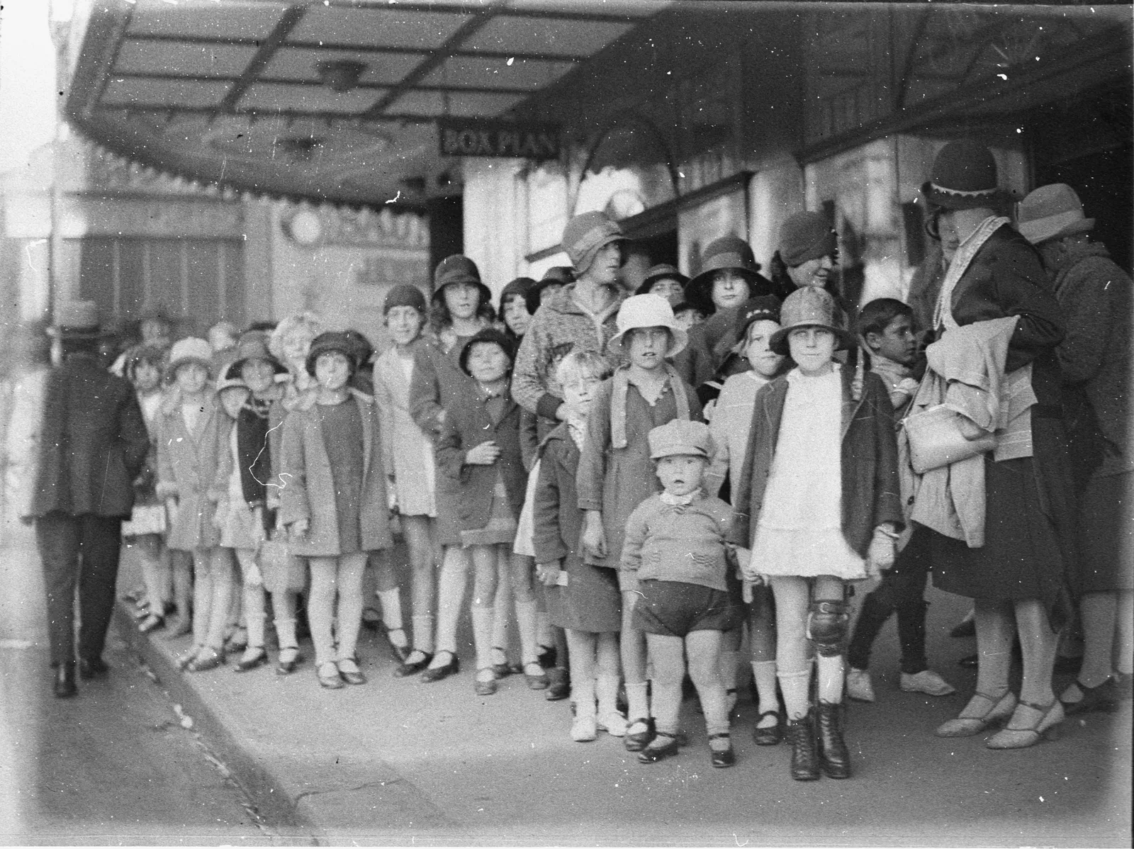 A crowd of children in cloche hats, felt hats and coats queues along the footpath under a picture theatre’s glass awning, with a few mothers and a toddler in a cap at the front.