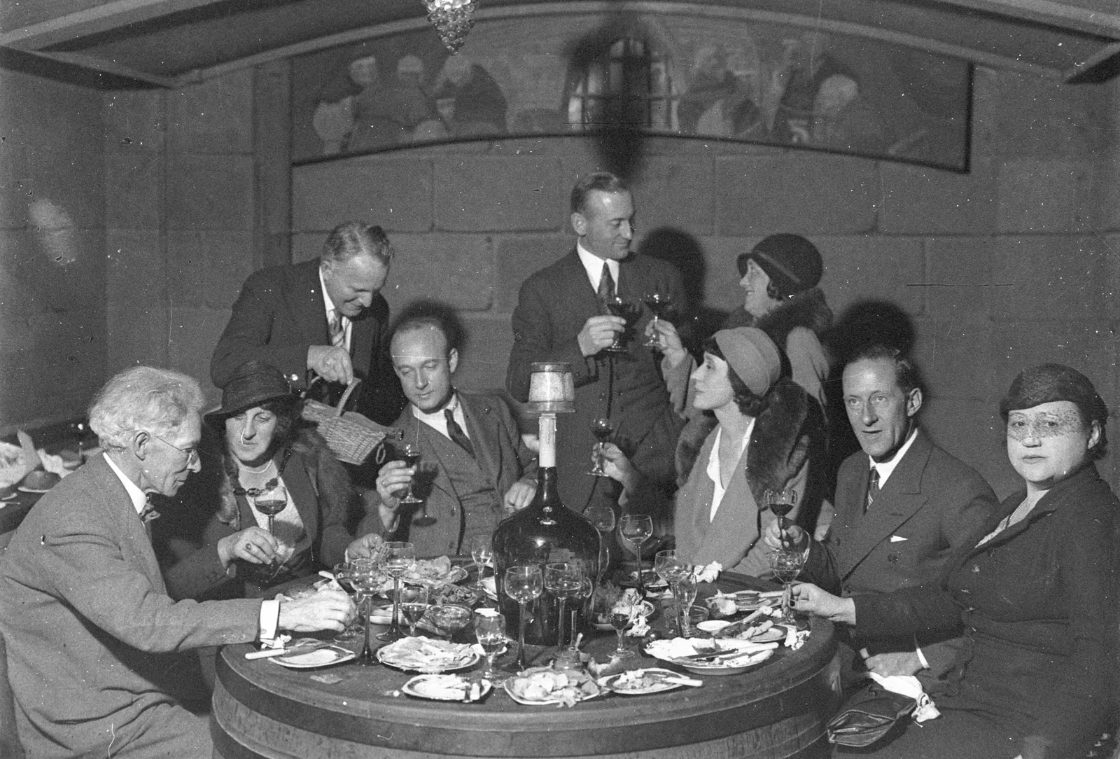 Leo Buring, standing at the left, pours from a wicker-cased bottle for eight guests round a barrel-top table in a vaulted cellar, the men in suits and the women in cloche hats raising their glasses under a mural of monks.